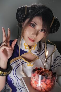  1girl 2026 blue_shirt bracelet brown_eyes brown_hair candle capcom chinese_(nationality) chinese_commentary chun-li chun-li_(cosplay) closed_mouth collared_shirt commentary cosplay cosplay_photo double_bun female_focus food fruit gold_trim hair_bun hair_ribbon hairdressing hand_up head_tilt highres jewelry jiaojiao_dingding_mao looking_at_viewer photo_(medium) pink_lips real_life ribbon shirt smile solo strawberry street_fighter street_fighter_6 two-tone_shirt upper_body v white_shirt yellow_ribbon 