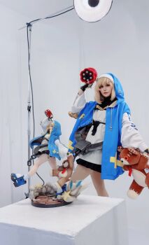 1boy 2025 androgyne_symbol androgynous aqua_eyes arc_system_works asian black_skirt blonde_hair blue_shoes blurry blurry_background blush brand_name_imitation bridget_(guilty_gear) bridget_(guilty_gear)_(cosplay) bush chain commentary cosplay cosplay_photo cross cuffs english_commentary figure figure_(medium) fingerless_gloves full_body gloves green_eyes grin guilty_gear guilty_gear_strive handcuffs headress highres holding holding_stuffed_toy holding_weapon hood hood_up hoodie indoors jacket japanese_(nationality) lips lipstick long_sleeves looking_at_viewer makeup male_focus medium_hair miniskirt one_eye_closed open_clothes open_hoodie photo_(medium) pleated_skirt puffy_long_sleeves puffy_sleeves real_life roger_(guilty_gear) shadow shoes skirt smile socks solo spread_fingers standing standing_on_one_leg stuffed_animal stuffed_toy teddy_bear teeth trap tree turtleneck upper_teeth_only walking weapon wink yo-yo yunomi_ochawan