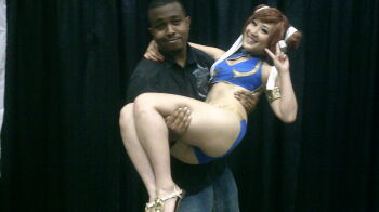 1boy 1girl adapted_costume armlet ass bikini breasts brown_hair capcom chubear chun-li chun-li_(cosplay) cosplay_photo dark-skinned_male dark_skin hair_buns holding jewelry looking_at_viewer navel photo_(medium) real_life sandals smile street_fighter swimsuit toes v