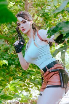 1girl argentine_(nationality) bare_arms black_gloves braid braided_ponytail breasts cleavage cosplay day fingerless_gloves gloves gun handgun highres holding holding_gun holding_weapon lara_croft lara_croft_(cosplay) lips medium_breasts shirt short_shorts shorts single_braid sophie_valentine tomb_raider tree weapon wide_hips