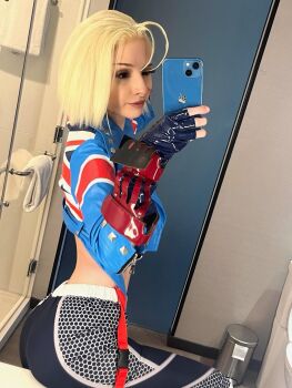 1girl ass black_gloves black_leggings blonde_hair blue_jacket cammy_white cammy_white_(cosplay) capcom cellphone cosplay cosplay_photo cropped_jacket female_focus fingerless_gloves gauntlets gloves highres holding holding_phone jacket leggings open_clothes open_jacket phone photo_(medium) real_life runabayo selfie short_hair smartphone solo street_fighter street_fighter_6 union_jack union_jack_print